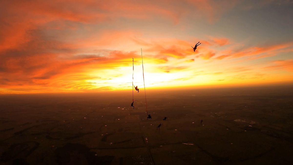 Skydivers silhouetted against a burning orange sunset above dark farmland